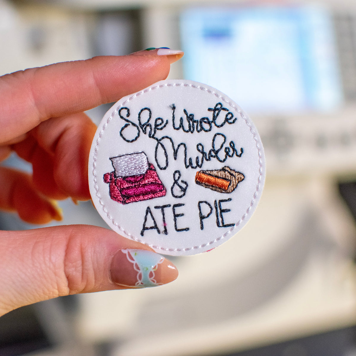 A hand holds a circular embroidered patch featuring the text 'She Wrote Murder & ATE PIE' with illustrations of a pink typewriter and an orange pie. The background shows a blurred sewing machine.