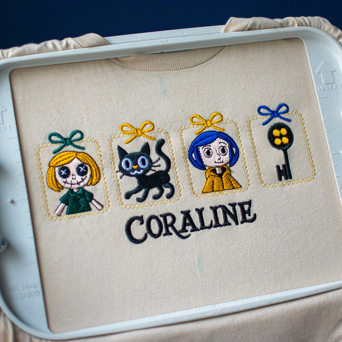 A close-up of a hand holding an embroidery hoop with a finished embroidered design. The design features four character panels with bows and the word 'CORALINE' stitched below in black.